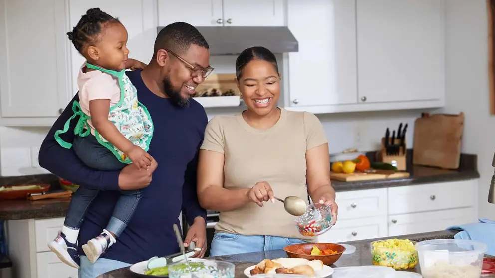 Une famille de trois est debout à côté d’un comptoir de cuisine; la mère prépare le repas et le père tient leur enfant d’un bras.