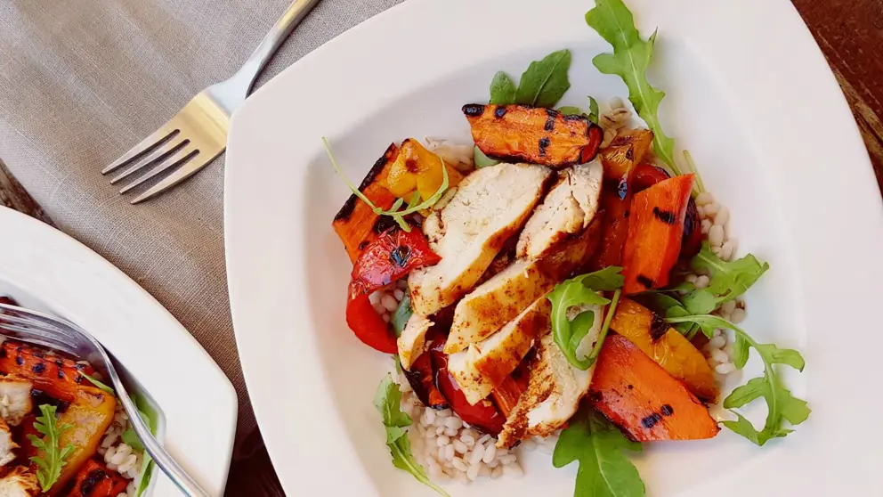 Poulet grillé sur lit d’orge dans une assiette blanche à côté de laquelle est posée une fourchette 