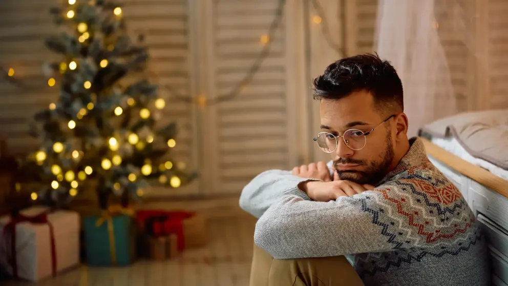 Un homme avec une expression triste est assis près du lit avec un sapin de Noël et des cadeaux en arrière-plan