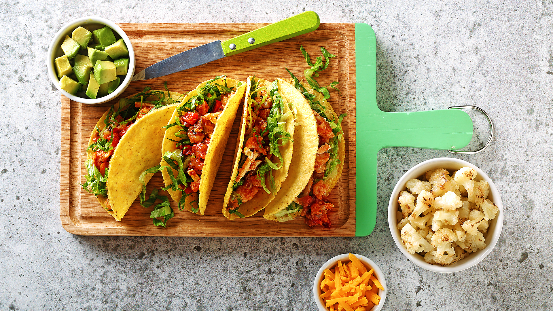 Tacos au chou-fleur présentés sur une planche à découper avec des bols de fromage, d’avocat et de chou-fleur.