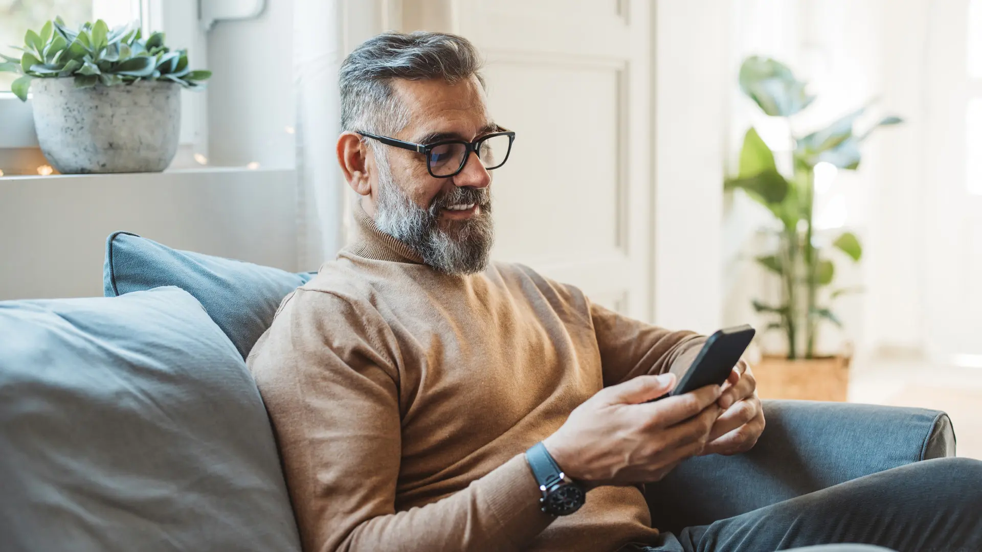 Un homme, assis sur un divan, regarde son téléphone intelligent.