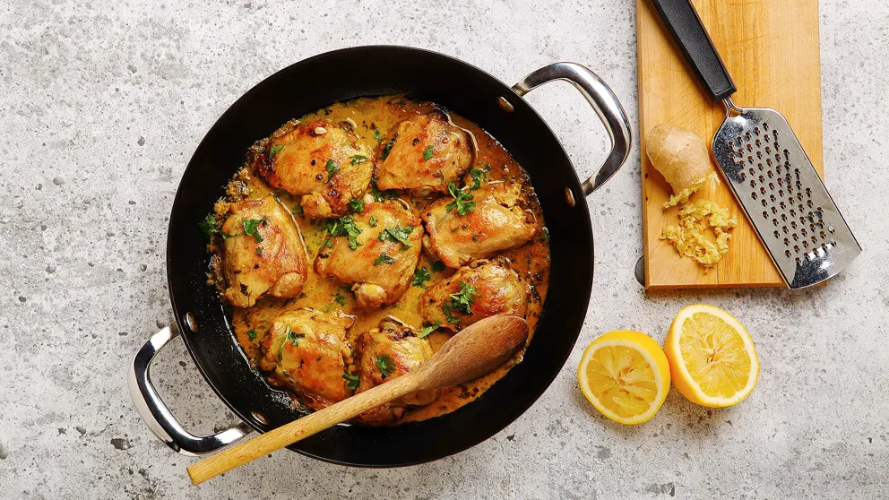 Poulet au cari, au citron et au gingembre dans une grande poêle, accompagné de gingembre râpé et d’un citron coupé en deux.