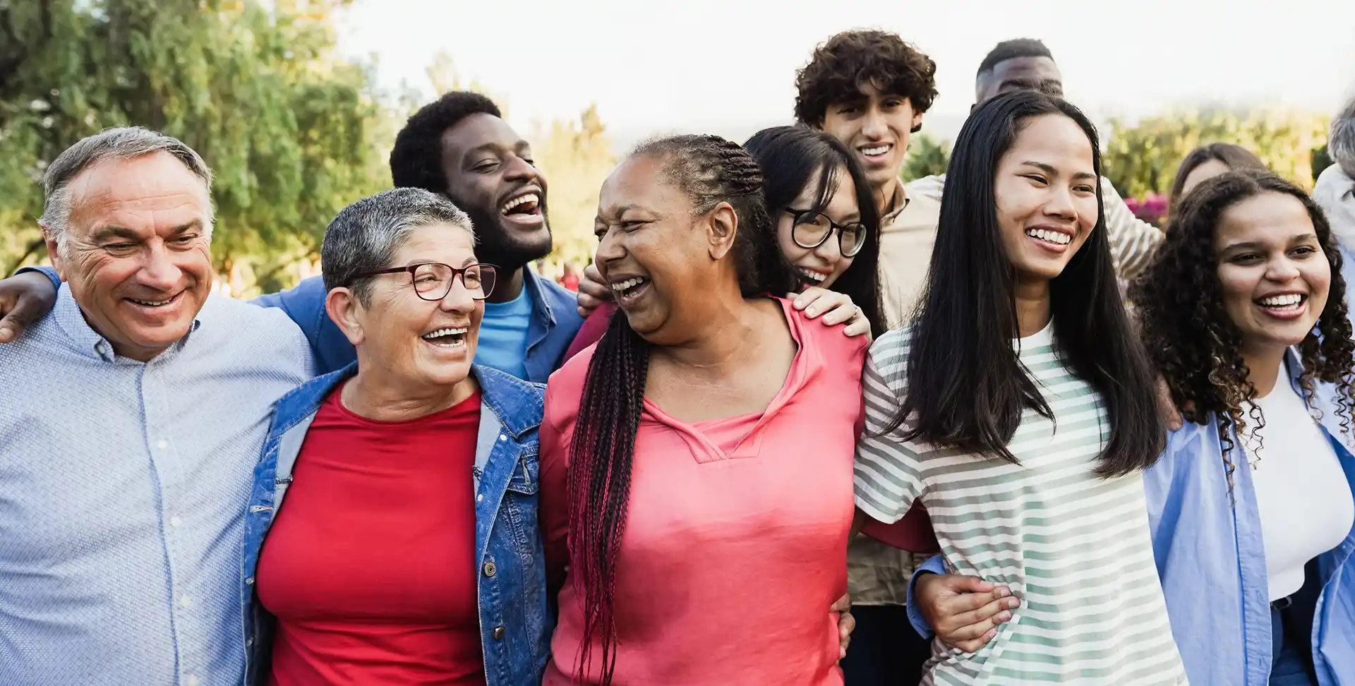 Un grand groupe diversifié de personnes s’enlacent à l’extérieur en souriant.