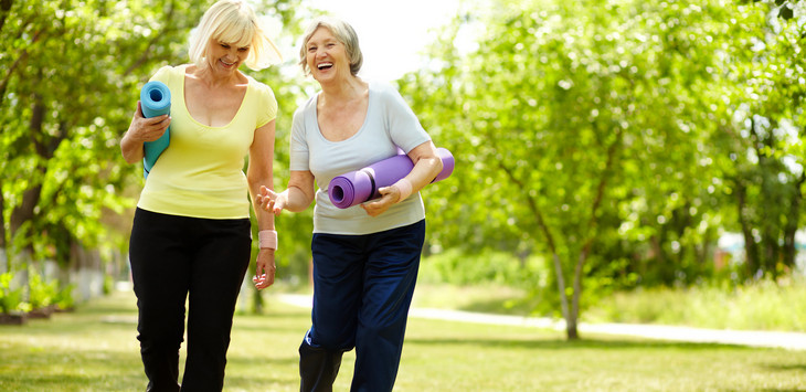 Two older women carrying yoga mats stroll through a park in the summertime, laughing. 